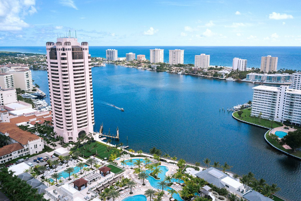 Aerial view of a coastal city with a canal, high-rise buildings, and a resort pool area in the foreground.