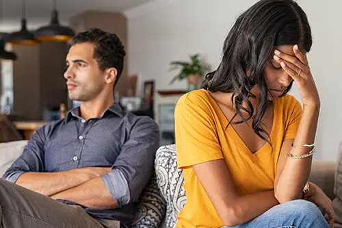 Couple sitting on a sofa facing away from each other; woman in a yellow top holds her head in distress, man in a blue shirt looks away with arms crossed.