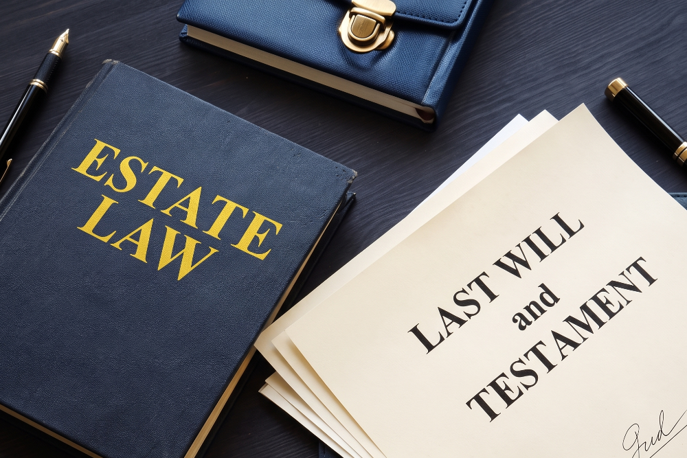 Dark blue 'Estate Law' book beside a stack of 'Last Will and Testament' documents on a dark wooden desk with pens nearby