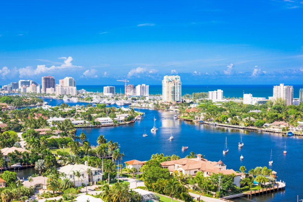 Sunny coastal city with a canal, boats, and palm-lined neighborhoods against a blue sea and skyline.