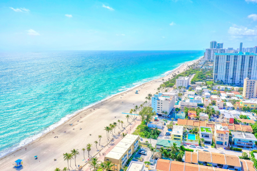 Aerial view of a sunny beach with turquoise water, palm trees, and a city skyline along the shore.