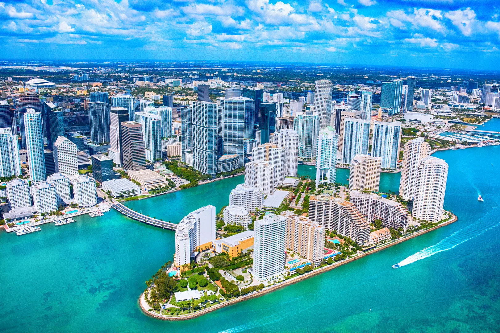 Aerial view of a dense waterfront city with many tall glass buildings along turquoise water and a curved bridge.