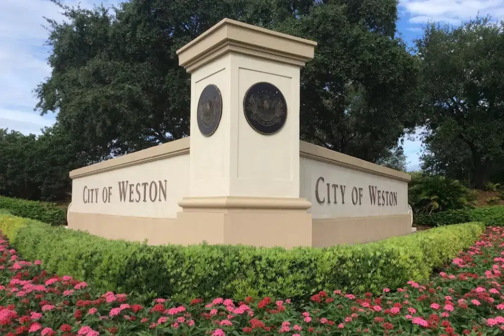 Entrance monument for the City of Weston with cream pillars and circular emblems, set among pink flowers and green hedges.