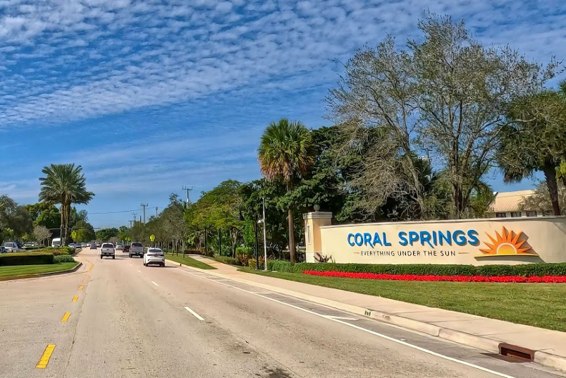 Entrance sign for Coral Springs along a palm-tree-lined street with a blue sky and a sun graphic; cars drive by in a suburban road scene.