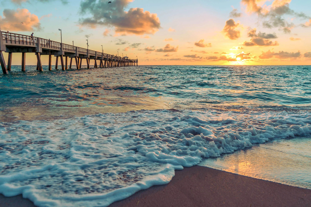 Dania Beach - Akilah Harris, PLLC. Sunset over the ocean with a wooden pier extending from the left into the water; foamy waves wash onto the sandy beach.