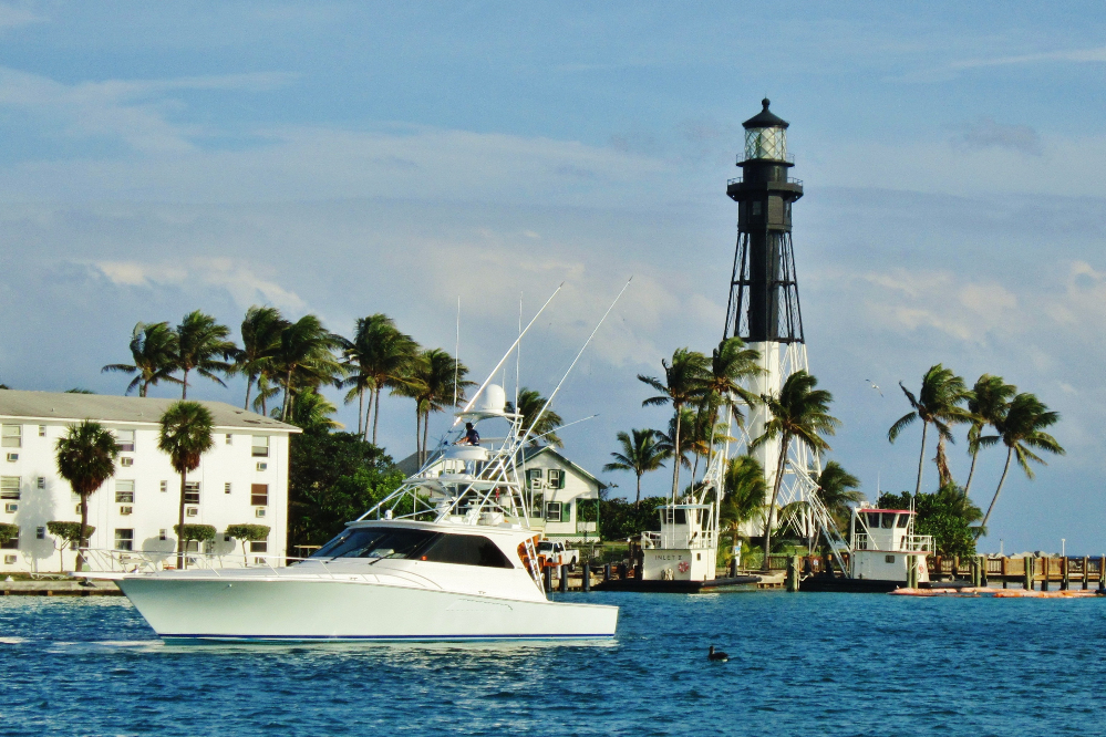 White motor yacht cruising near a shoreline with palm trees and a tall lighthouse in the background.