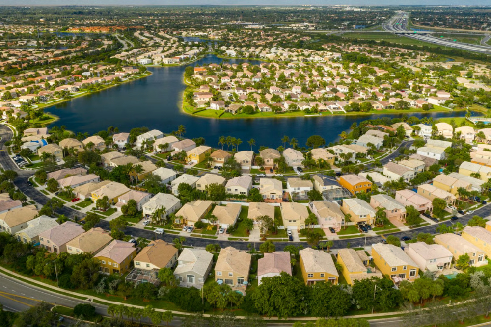 Aerial view of a suburban neighborhood circling a large lake with rows of beige and pastel houses along curving streets.
