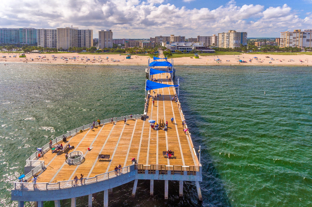 Aerial view of a long wooden pier with blue shade canopies extending into green-blue water toward a sandy beach and high-rise hotels in the distance, with people strolling.