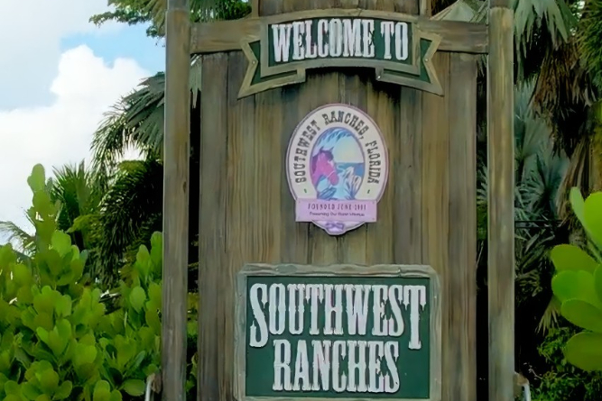 Wooden welcome sign for Southwest Ranches, with a circular emblem and tropical plants surrounding it.