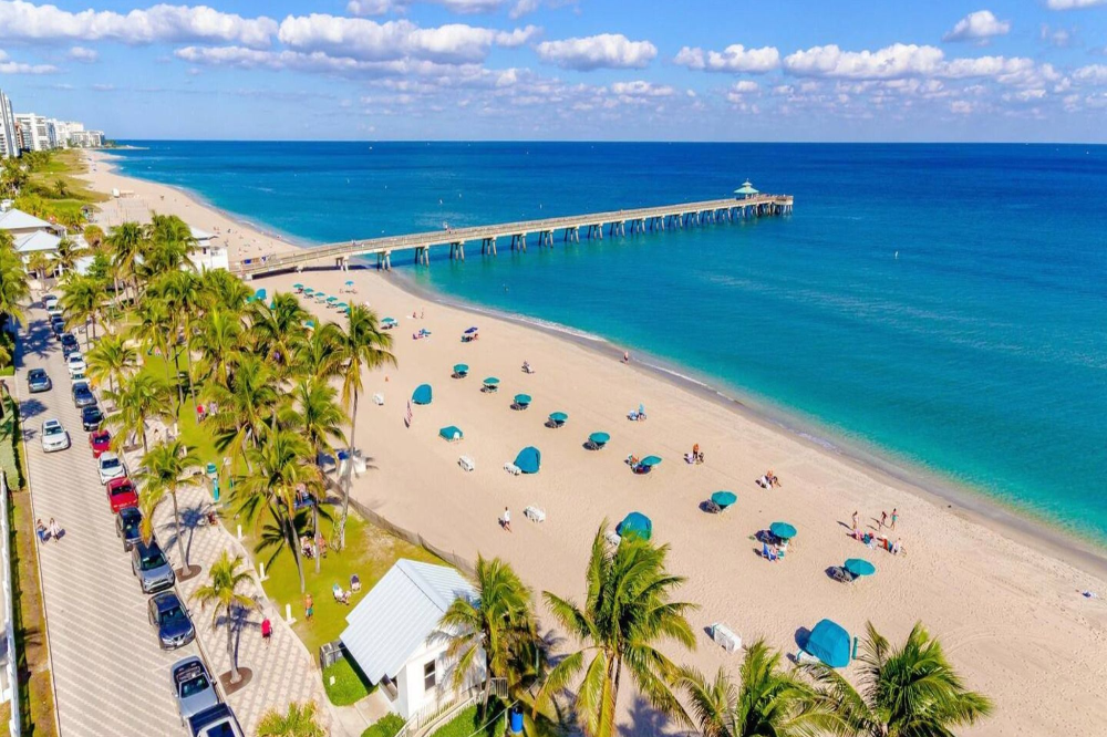 Deerfield Beach - Akilah Harris, PLLC. Aerial view of a sunny beach with turquoise water, palm trees, blue umbrellas, and a long wooden pier extending into the ocean.