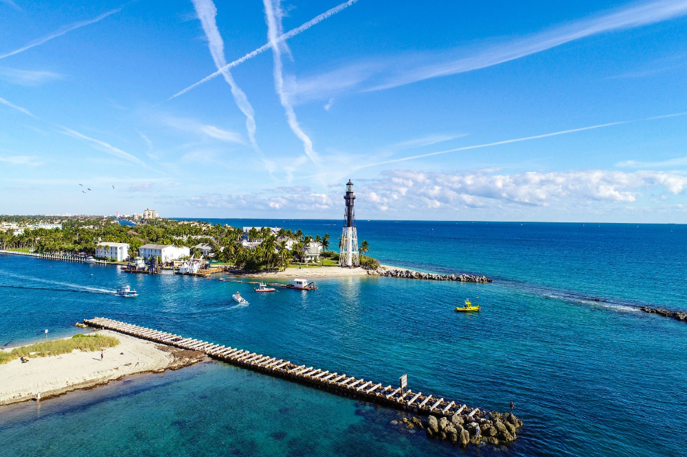 Lighthouse on a small rocky island with palm trees, blue sea, and a long jetty extending into calm water with boats nearby.