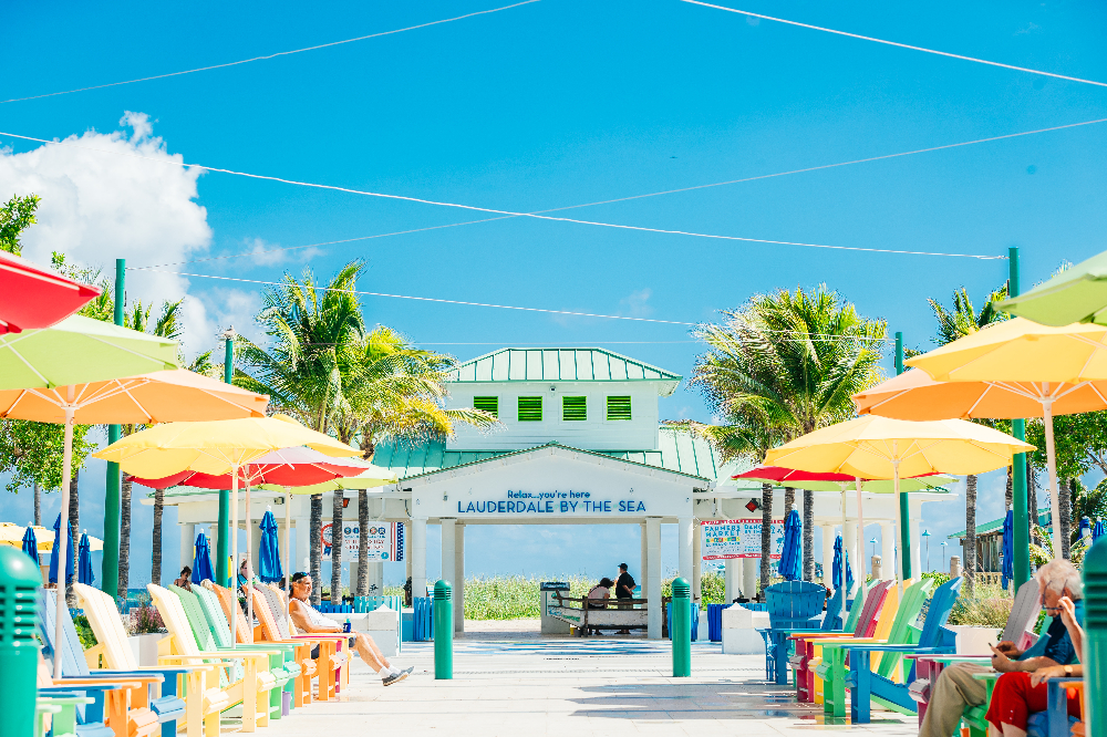 Lauderdale By the Sea - Akilah Harris, PLLC. Colorful beachside boardwalk with bright chairs and umbrellas leading to a white pier in Lauderdale-by-the-Sea under a blue sky.