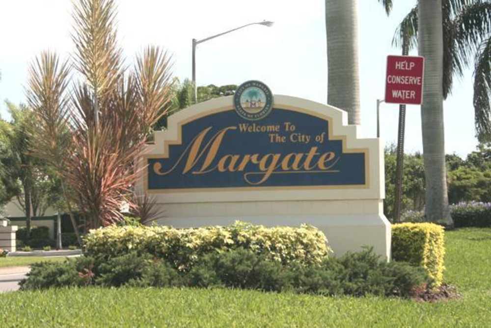 Welcome to the City of Margate sign with palm trees and greenery in front of landscaping; red 'Help Conserve Water' sign nearby.