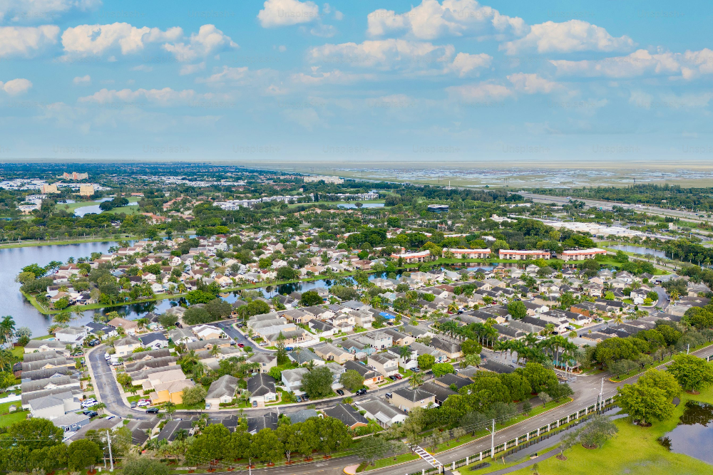 Aerial view of a dense suburban neighborhood with winding streets, rows of houses, trees, and surrounding canals under a blue sky with clouds.