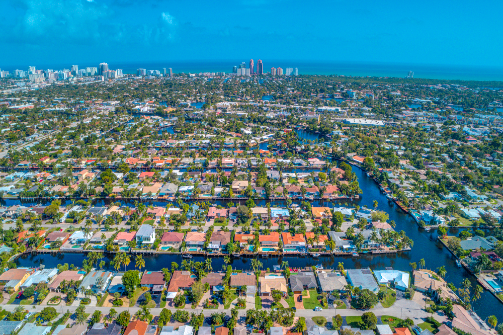 Aerial view of a coastal city with canals weaving through a residential neighborhood and palm trees toward the ocean skyline.