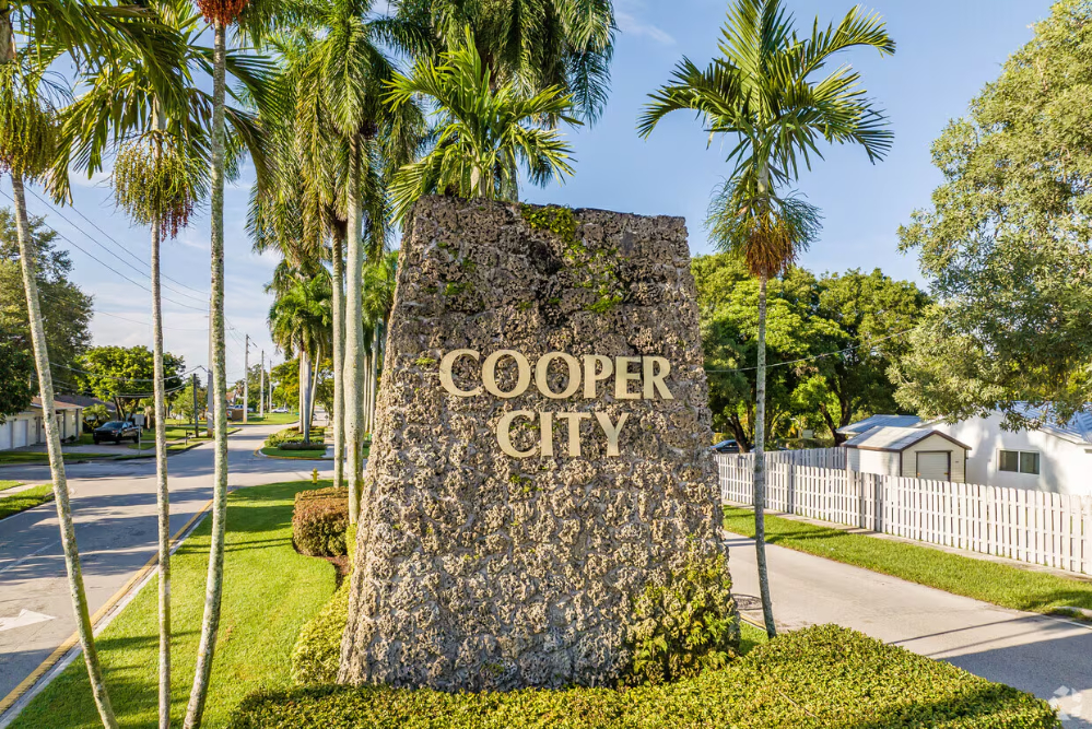 cooper-city-fl - Akilah Harris, PLLC. Rocky monument with the words COOPER CITY, flanked by tall palm trees along a sunny suburban street entrance.