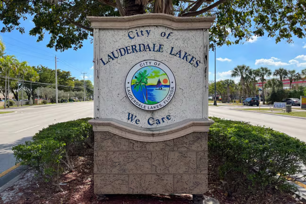 Stone city sign reading 'City of Lauderdale Lakes' with a circular Florida-themed logo; motto 'We Care' below, set against a blue sky and palm-lined street in Lauderdale Lakes, FL.