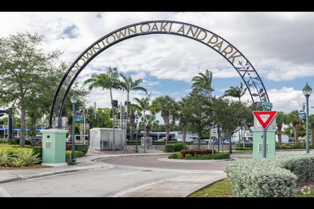 Entrance to a park with an ornate metal arch reading 'Downtown Oakland Park' over a circular driveway and palm trees in the background.