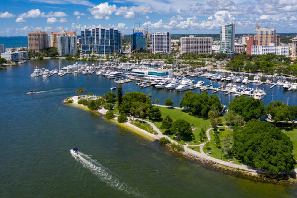 Parkland - Akilah Harris, PLLC. Aerial view of a marina with many white yachts docked, blue water, and a green park beside the city skyline.