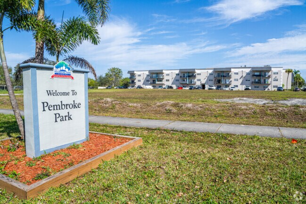 Pembroke Park - Akilah Harris, PLLC. Pembroke Park welcome sign with palm trees and a row of white apartment buildings in the distance.